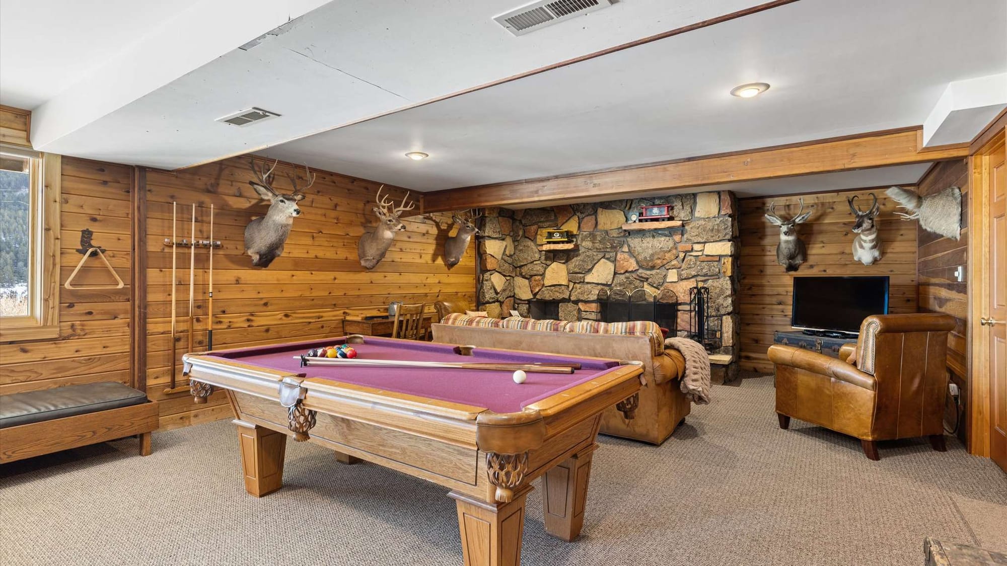 Rustic game room with wood-paneled walls, mounted deer heads, a stone fireplace, and a pool table, complemented by cozy leather seating and a TV lounge area.