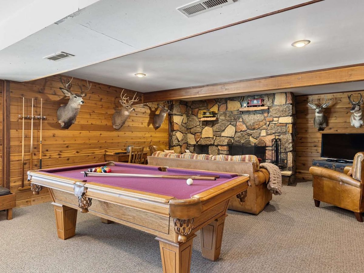 Rustic game room with wood-paneled walls, mounted deer heads, a stone fireplace, and a pool table, complemented by cozy leather seating and a TV lounge area.