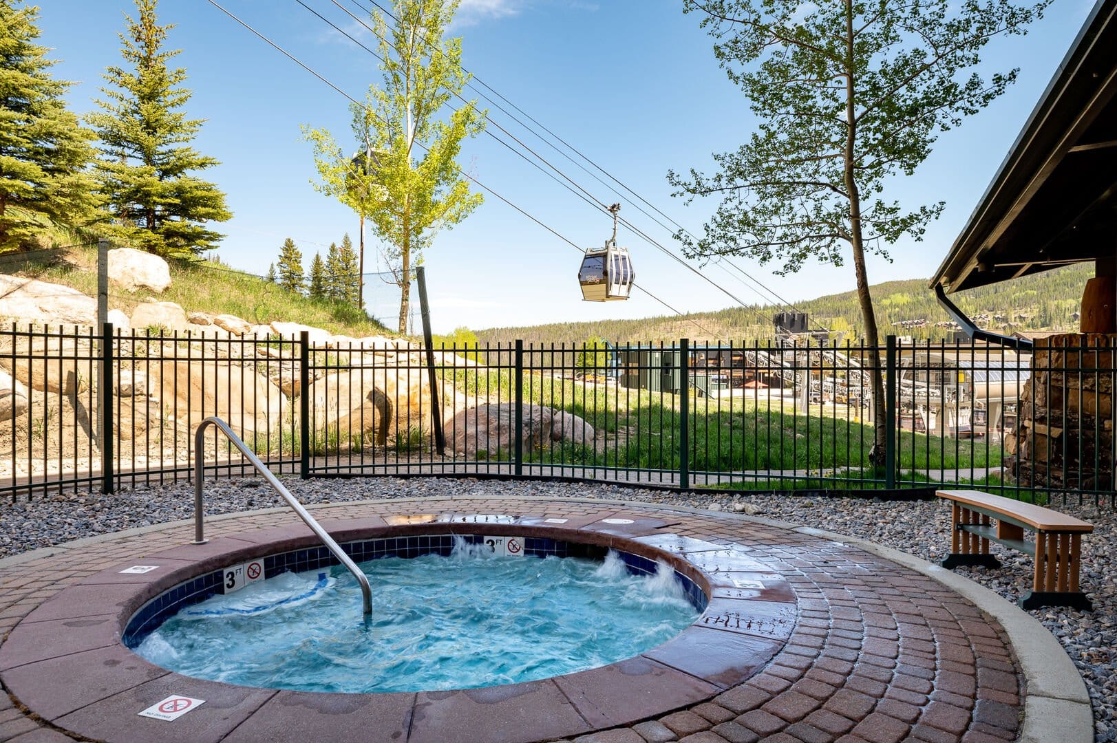 An outdoor hot tub surrounded by stone pavers and a black metal fence, with gondola lifts passing overhead and scenic mountain views in the background on a sunny day.