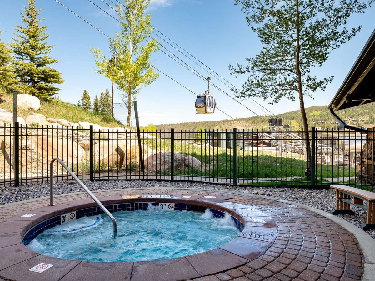 An outdoor hot tub surrounded by stone pavers and a black metal fence, with gondola lifts passing overhead and scenic mountain views in the background on a sunny day.