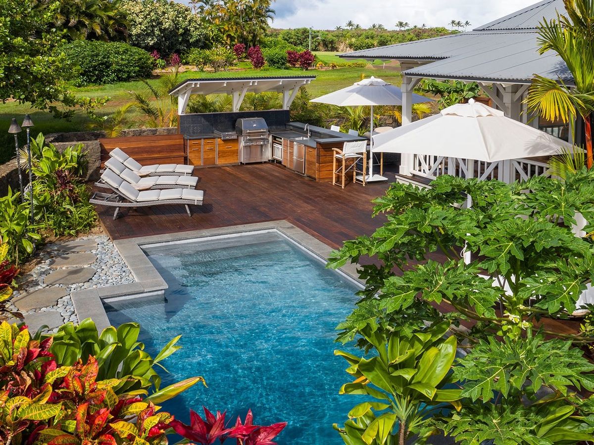 This tropical outdoor space features a private pool surrounded by bright island plants and a beautiful wood deck. The open-air kitchen and lounge chairs make it easy to relax, cook, and enjoy long sunny days with your family. It feels like a peaceful hideaway where every moment becomes a memory.
