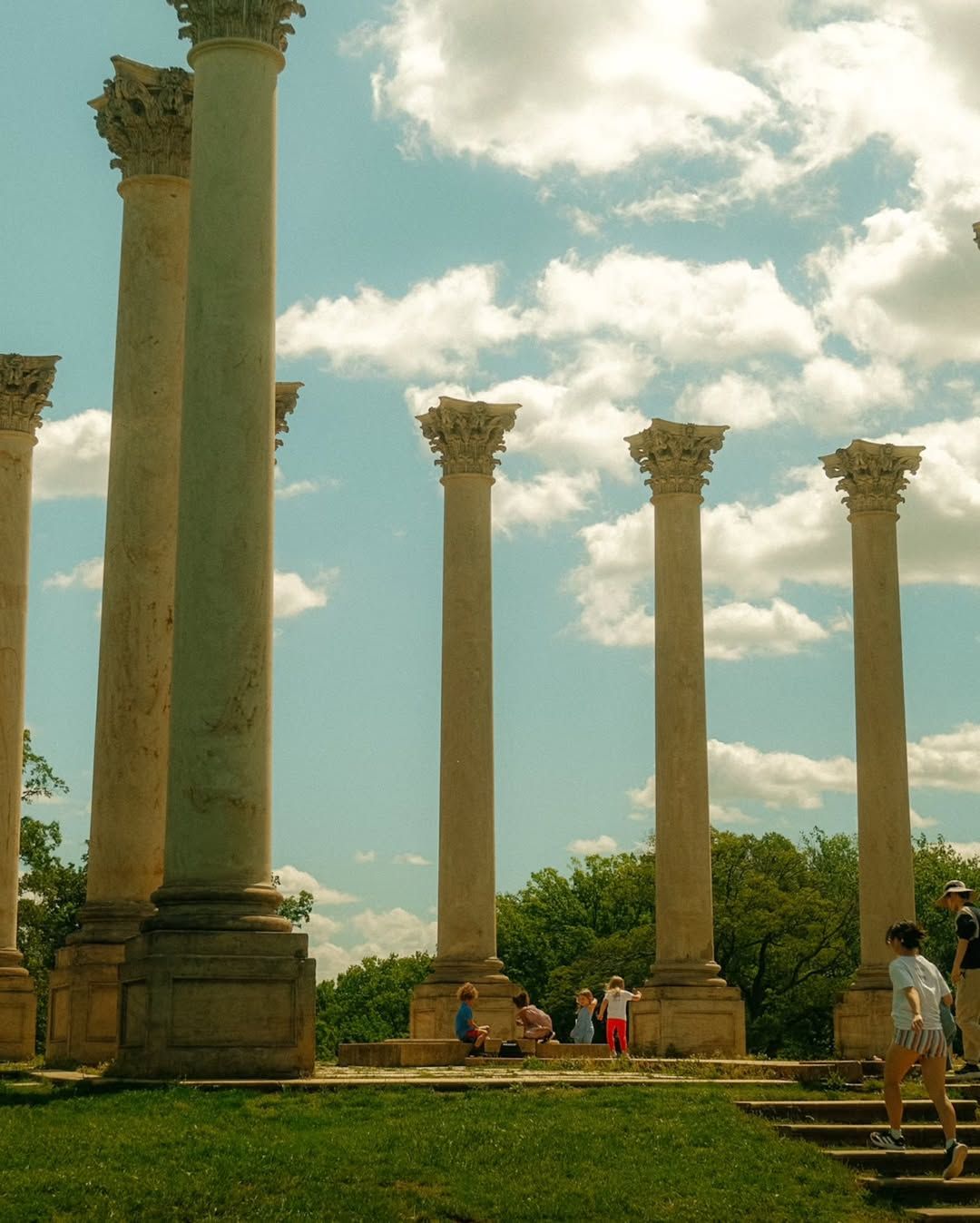Visitors explore the iconic Capitol Columns at the US National Arboretum in Washington DC, where a row of towering Corinthian columns salvaged from the original Capitol building rise dramatically against a blue sky with billowing clouds. 