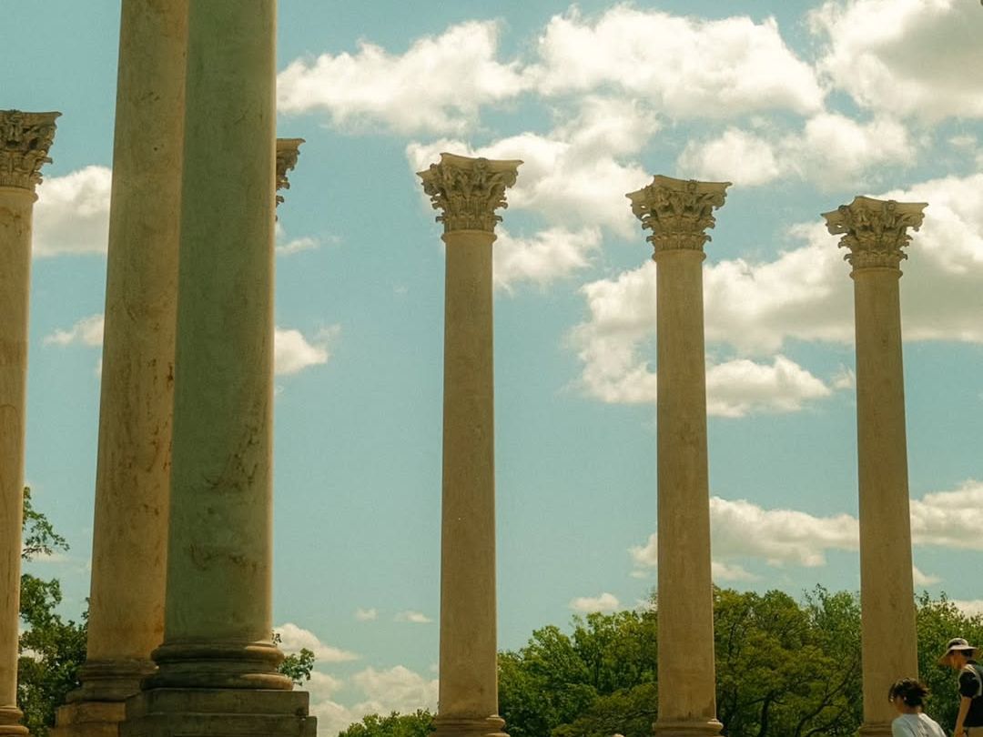Visitors explore the iconic Capitol Columns at the US National Arboretum in Washington DC, where a row of towering Corinthian columns salvaged from the original Capitol building rise dramatically against a blue sky with billowing clouds.
