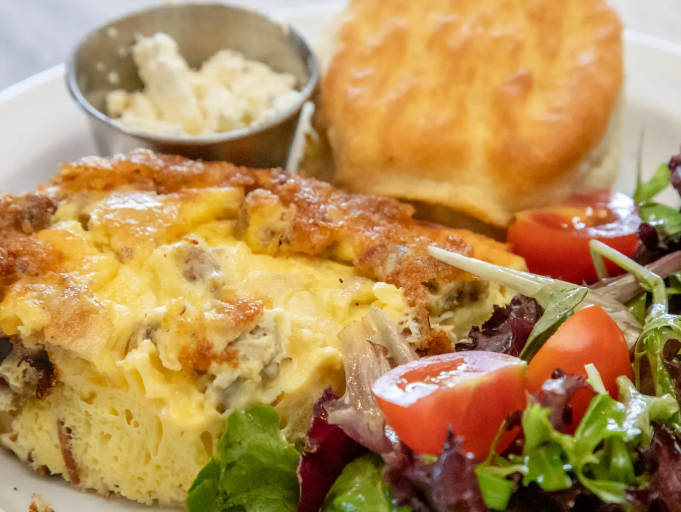 A thick, golden slice of baked egg casserole topped with melted cheese and sausage crumbles, served with a fresh mixed-greens salad, cherry tomatoes, a fluffy biscuit, and a side of whipped butter.