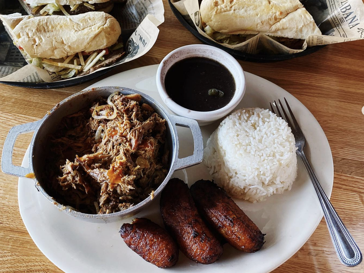 A plate of Cuban food featuring shredded ropa vieja in a small pot, white rice, sweet plantains, and a side of black beans, with Cuban sandwiches in the background served on newspaper-style paper.