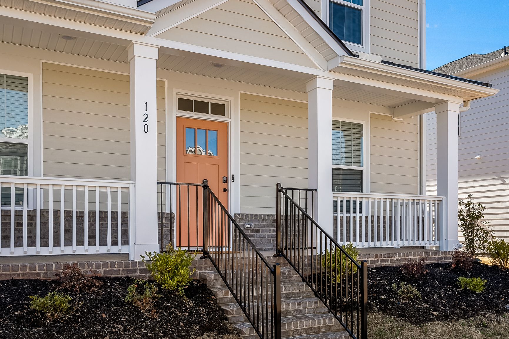 Front view of a beige house with a brick foundation, white trim, a coral-colored front door, black railings, and a small covered porch with white railing.