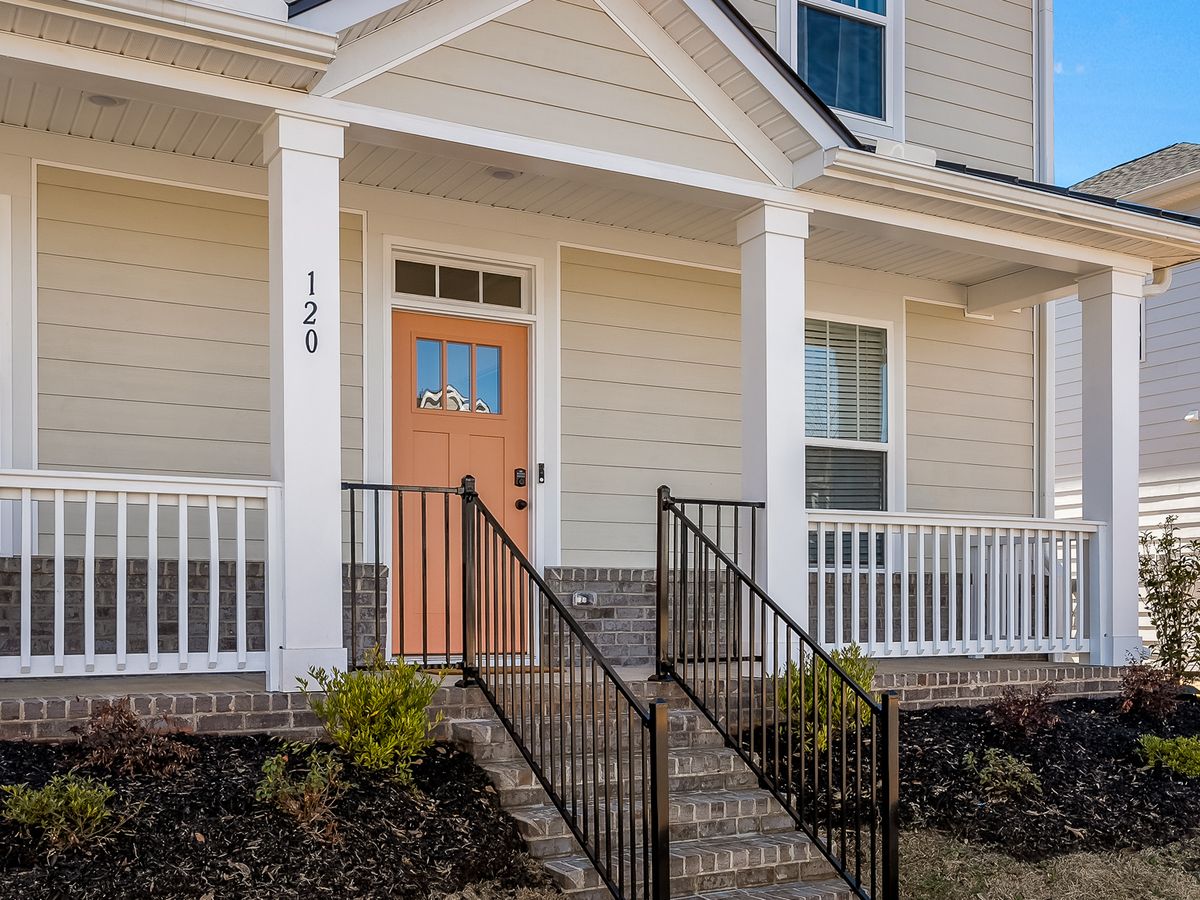 Front view of a beige house with a brick foundation, white trim, a coral-colored front door, black railings, and a small covered porch with white railing.