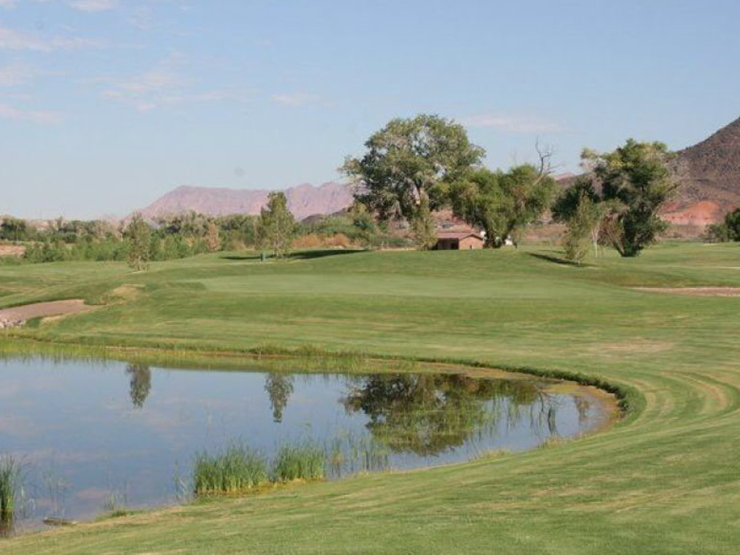 A serene golf course scene featuring a reflective water hazard, lush green fairways, mature trees, and a desert mountain backdrop under a clear blue sky