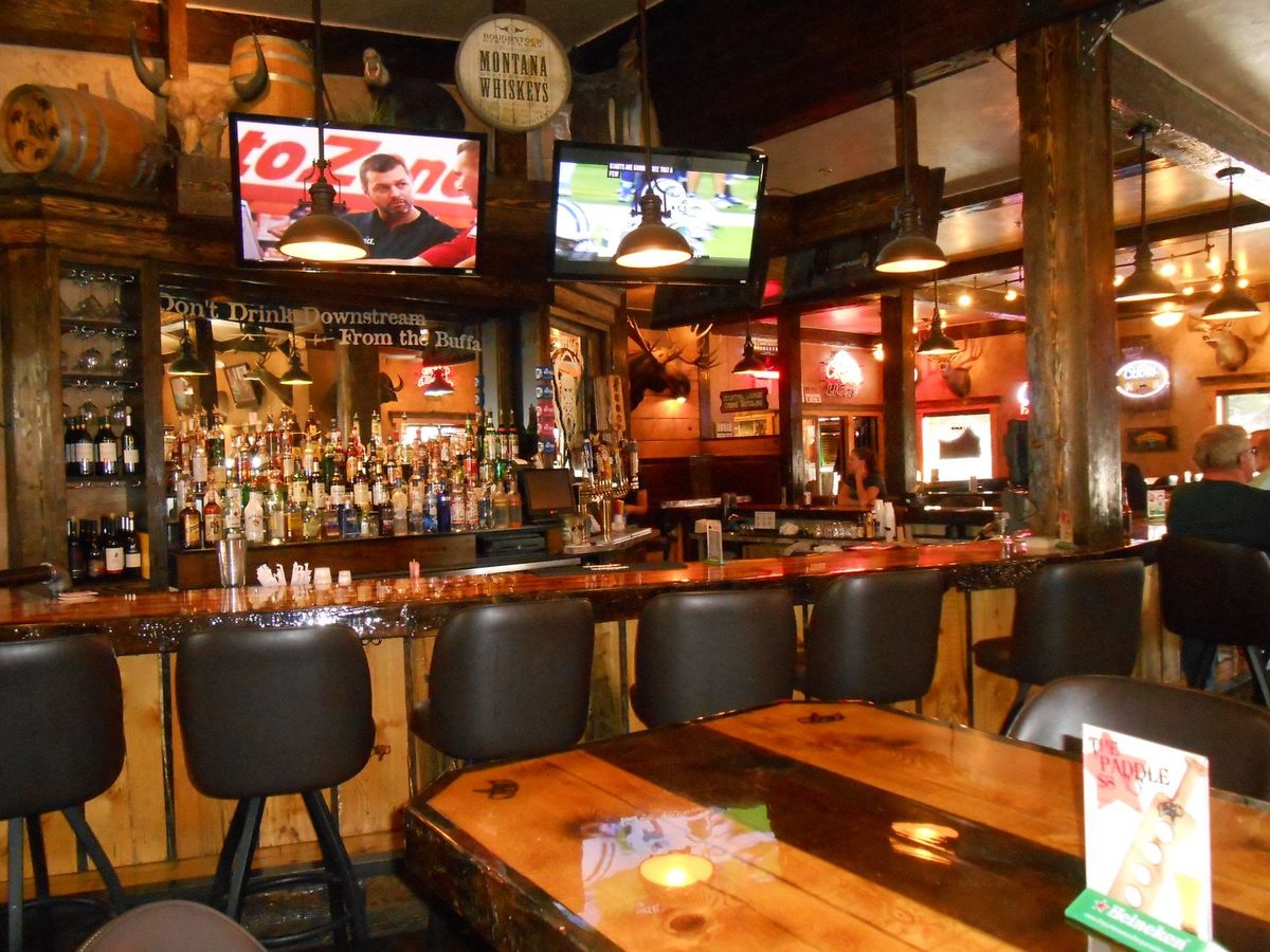 The rustic wooden bar at The Buffalo Bar in West Yellowstone features whiskey bottles, mounted decor, and sports playing on TV screens. Warm lighting gives it a true mountain-town feel. It’s a popular place for drinks and comfort food near Yellowstone National Park.
