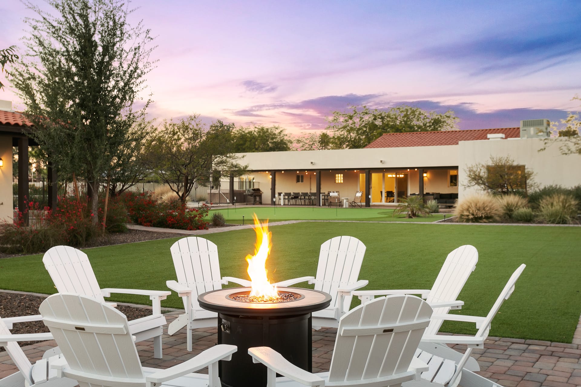  Backyard patio at sunset with a central fire pit surrounded by white Adirondack chairs, overlooking a landscaped lawn and a modern desert-style home with covered outdoor seating.