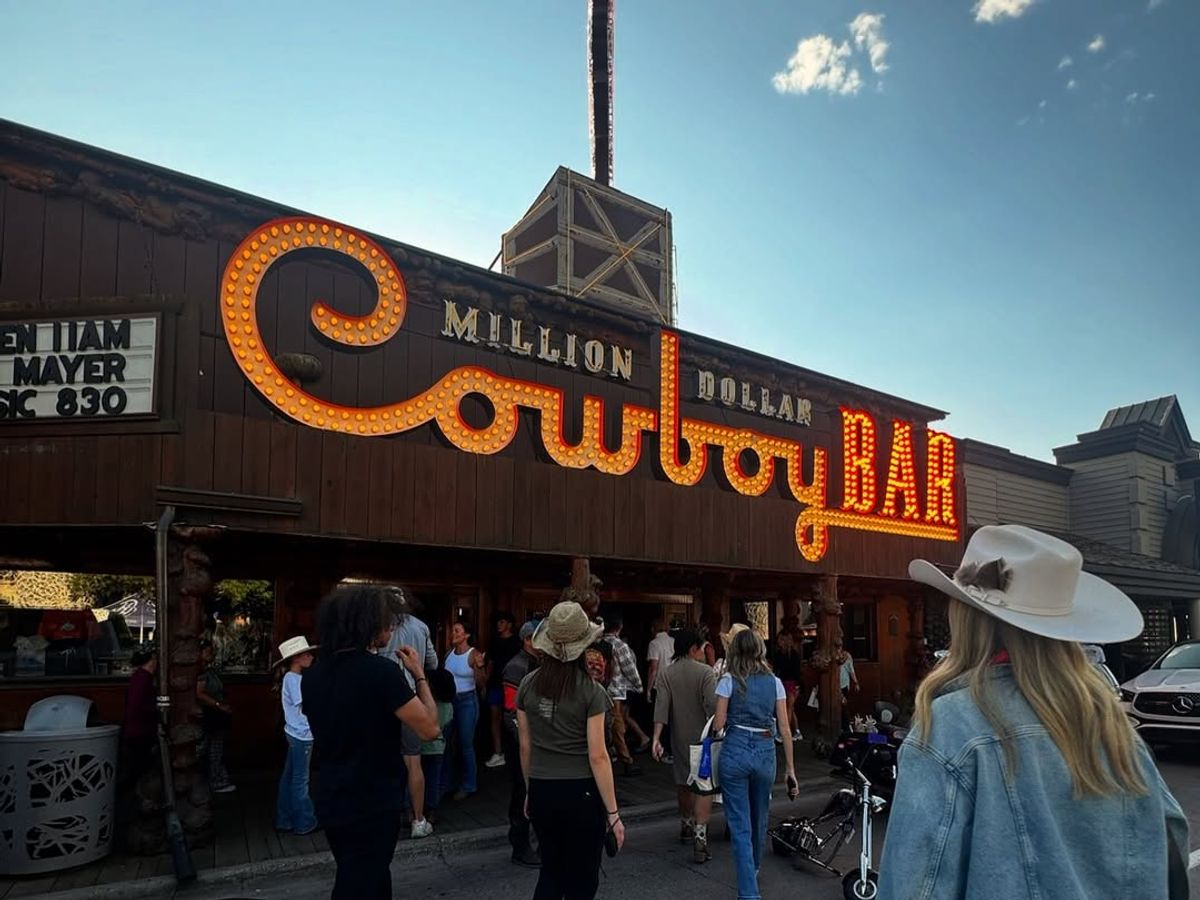  The iconic glowing marquee of the Million Dollar Cowboy Bar lights up its rustic log facade as a crowd of visitors gathers on the sidewalk below. A woman in a cream cowboy hat and denim jacket stands in the foreground beneath a bright blue evening sky.