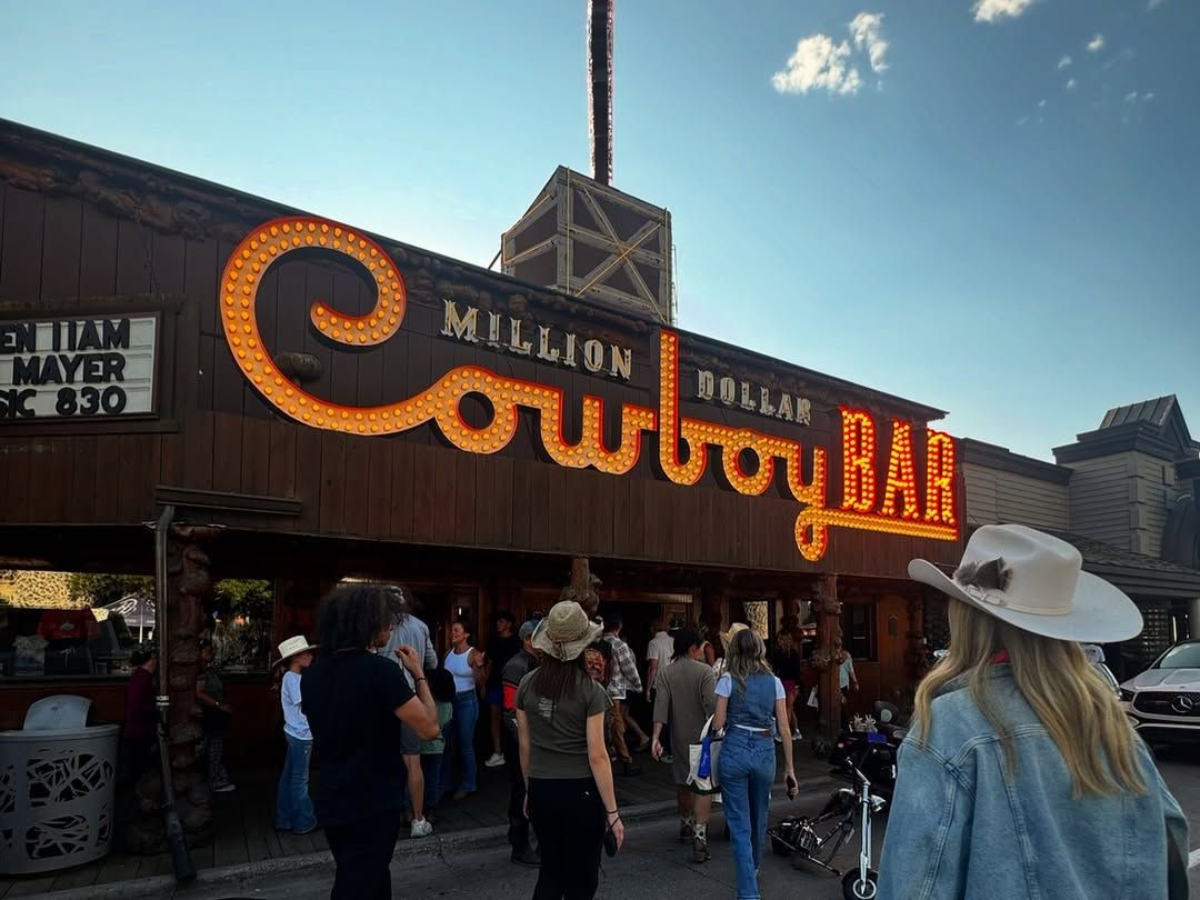 The iconic glowing marquee of the Million Dollar Cowboy Bar lights up its rustic log facade as a crowd of visitors gathers on the sidewalk below. A woman in a cream cowboy hat and denim jacket stands in the foreground beneath a bright blue evening sky.