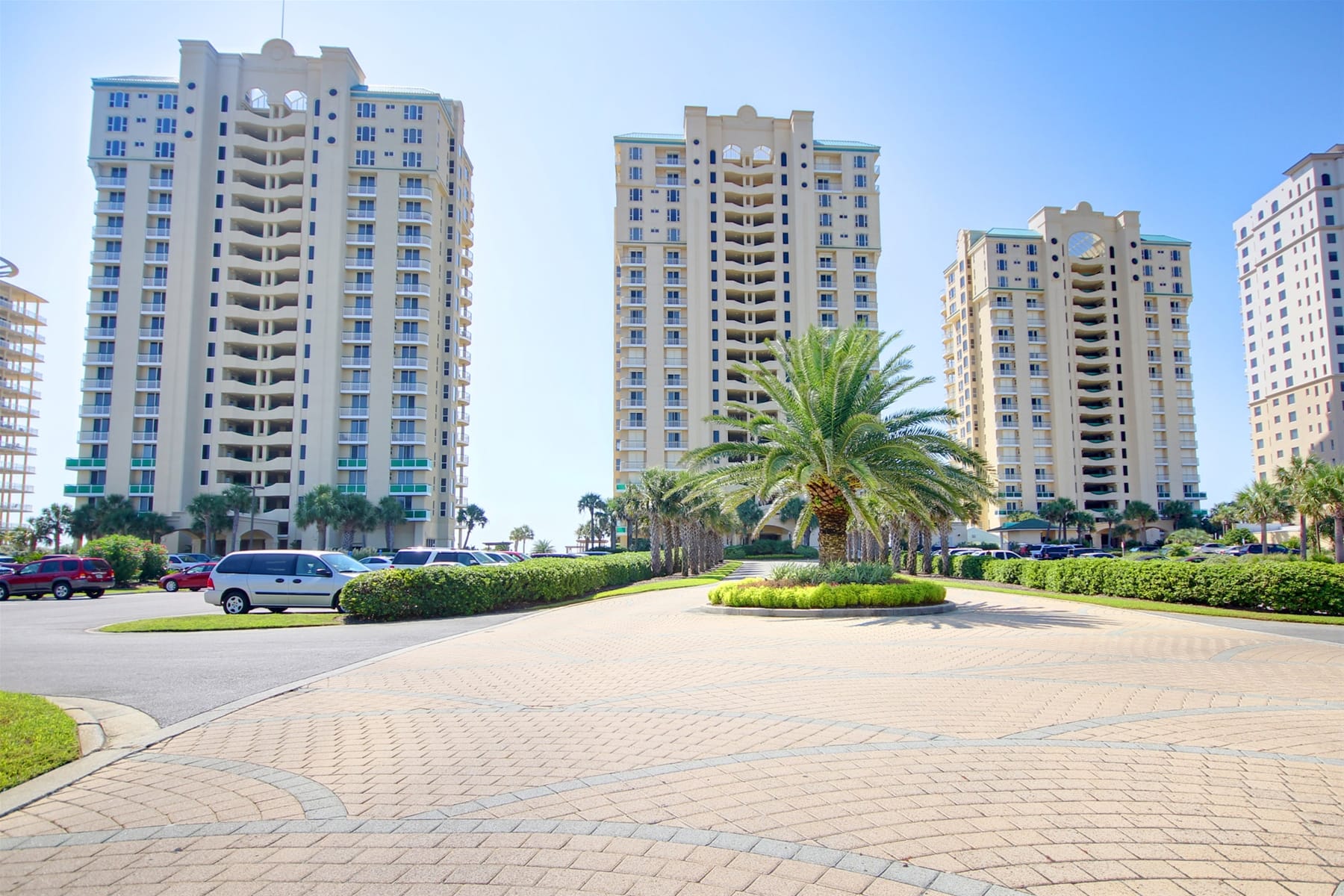 High-rise beachfront condos stand tall in Perdido Key, Florida, surrounded by palm trees and a sunny coastal skyline. The entrance drive features landscaped greenery and wide open space near the Gulf of Mexico. This beach destination is known for white sand shores and relaxing Florida vacations.