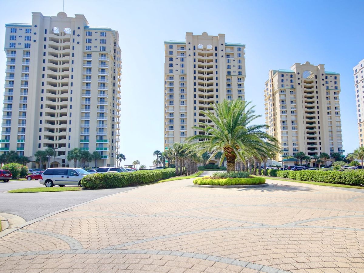 High-rise beachfront condos stand tall in Perdido Key, Florida, surrounded by palm trees and a sunny coastal skyline. The entrance drive features landscaped greenery and wide open space near the Gulf of Mexico. This beach destination is known for white sand shores and relaxing Florida vacations.