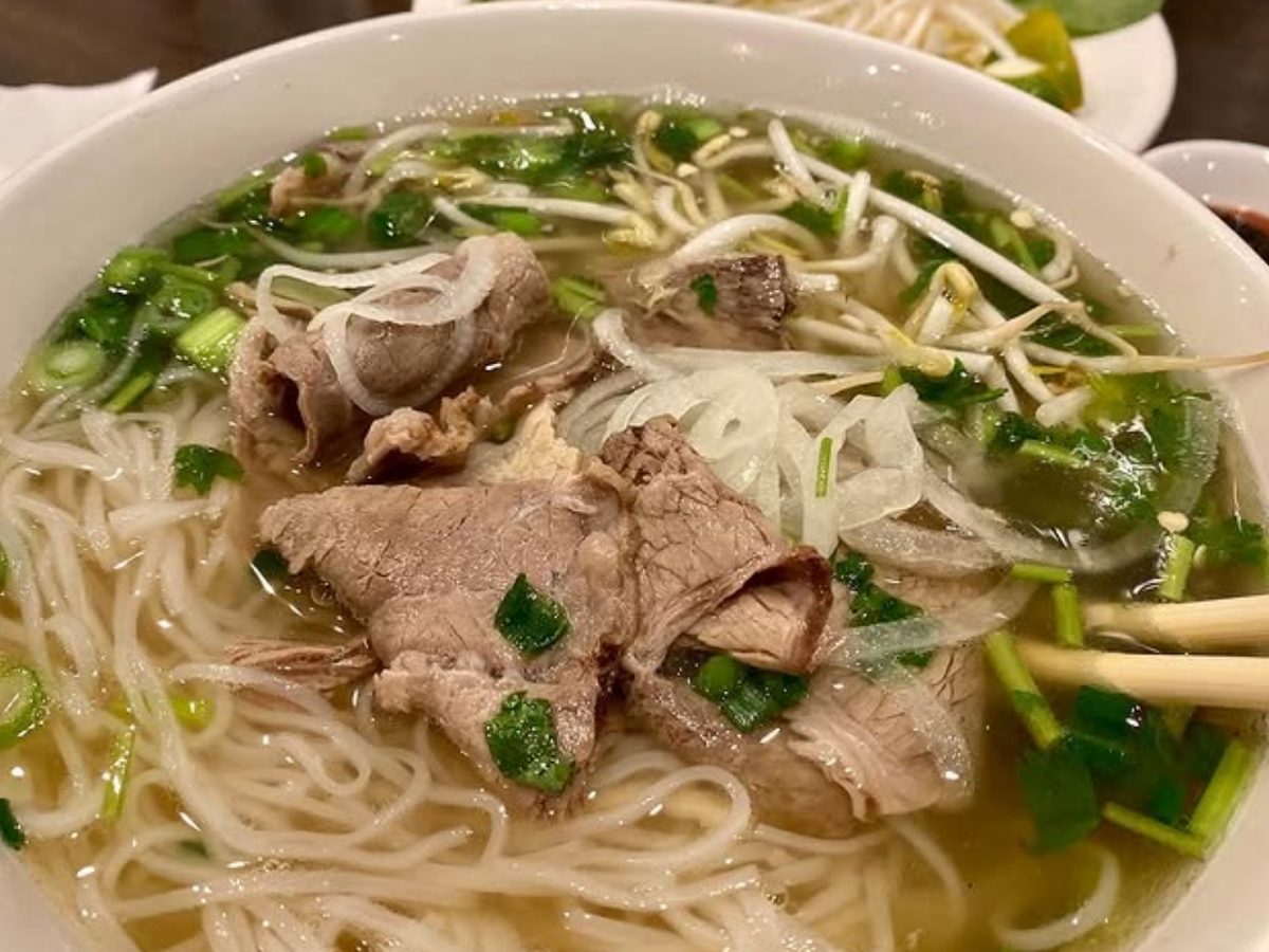A steaming bowl of Vietnamese pho with rice noodles, sliced beef, bean sprouts, onions, and green herbs in a clear broth, served with chopsticks.