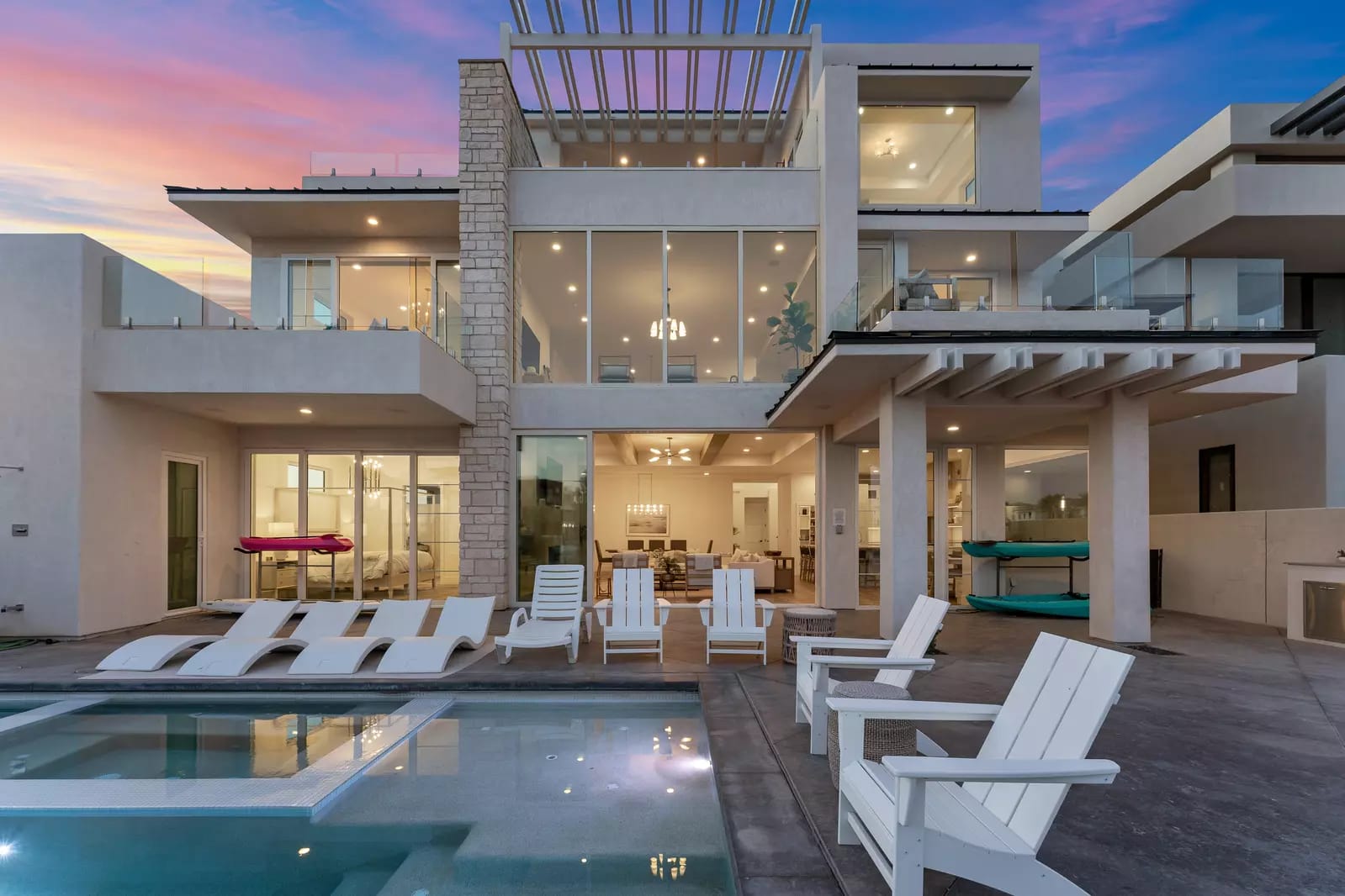 Contemporary multi-level home with expansive glass walls, multiple balconies, and a poolside patio set against a sunset sky.