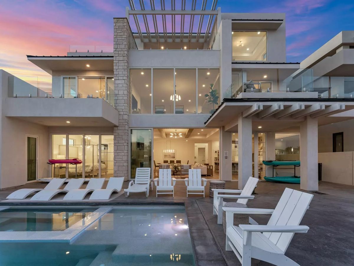 Contemporary multi-level home with expansive glass walls, multiple balconies, and a poolside patio set against a sunset sky.