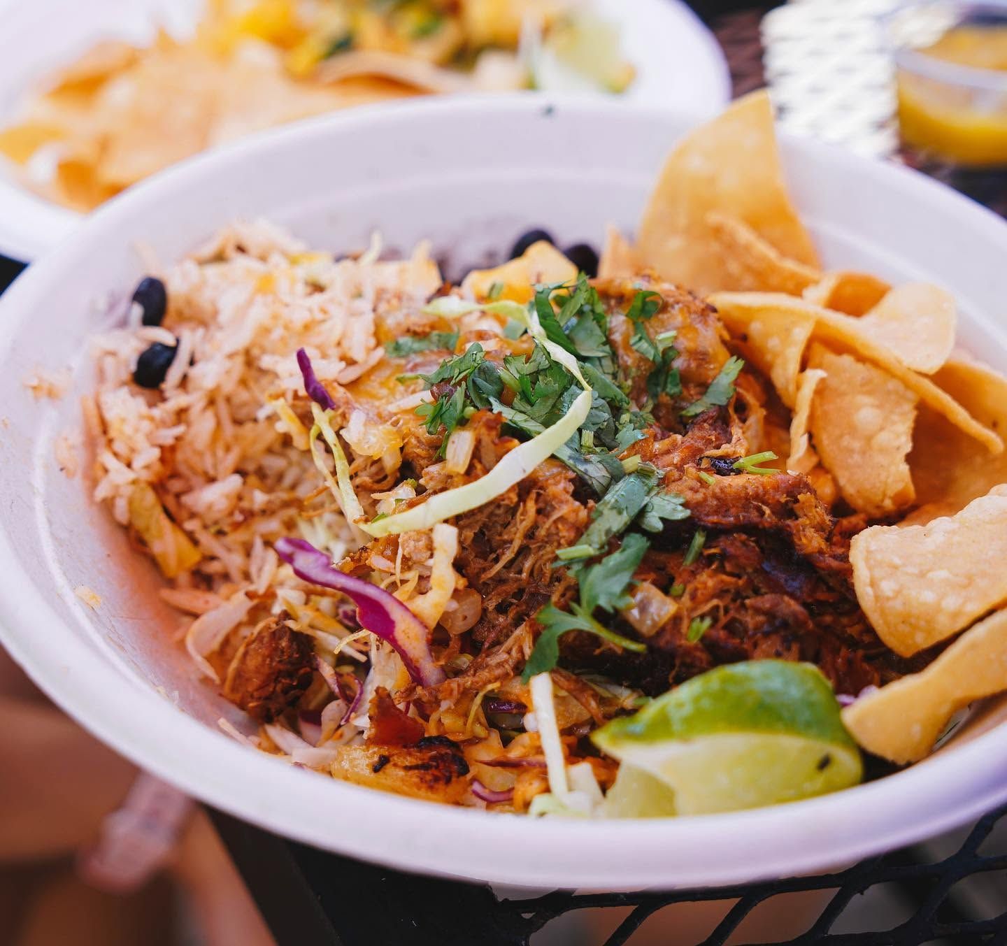  generous burrito bowl brims with seasoned rice, black beans, shredded slaw, tender carnitas, melted cheese, and fresh cilantro, garnished with a lime wedge and crisp tortilla chips. Additional plates of food sit in the background on a sunny patio table.