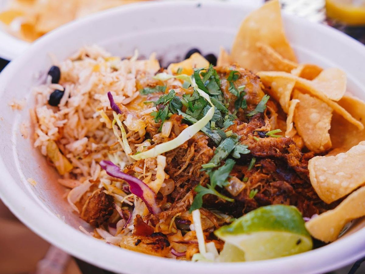 generous burrito bowl brims with seasoned rice, black beans, shredded slaw, tender carnitas, melted cheese, and fresh cilantro, garnished with a lime wedge and crisp tortilla chips. Additional plates of food sit in the background on a sunny patio table.