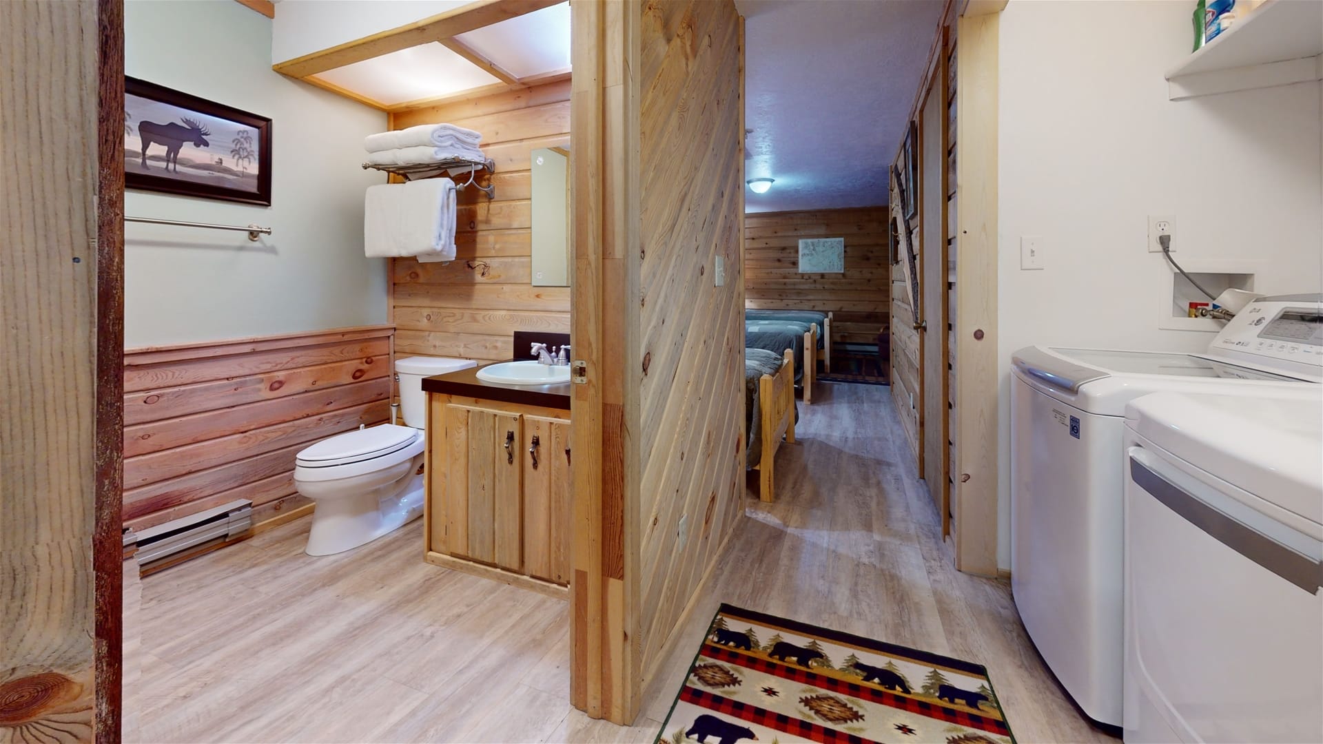 Cabin-style hallway with a bathroom, laundry area, and a view into a bedroom with wooden walls and rustic decor.