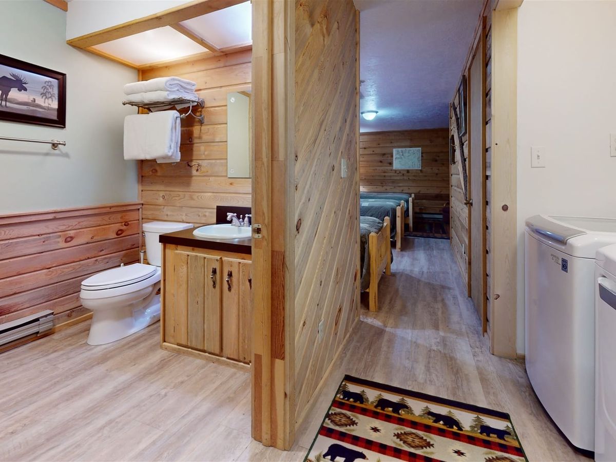 Cabin-style hallway with a bathroom, laundry area, and a view into a bedroom with wooden walls and rustic decor.