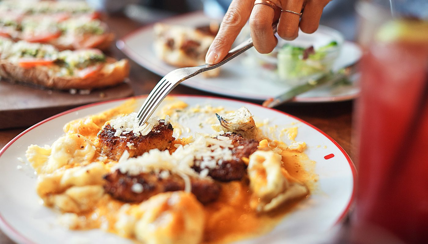 A close-up shot shows someone cutting into a creamy pasta dish topped with crispy chicken and melted cheese. The food looks rich, warm, and comforting—like a homemade favorite. It captures the farm-fresh, hearty style Founding Farmers is known for.