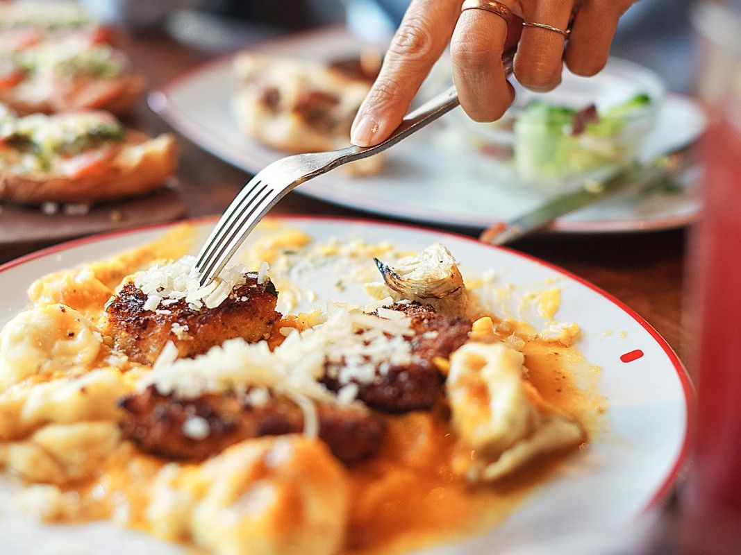 A close-up shot shows someone cutting into a creamy pasta dish topped with crispy chicken and melted cheese. The food looks rich, warm, and comforting—like a homemade favorite. It captures the farm-fresh, hearty style Founding Farmers is known for.