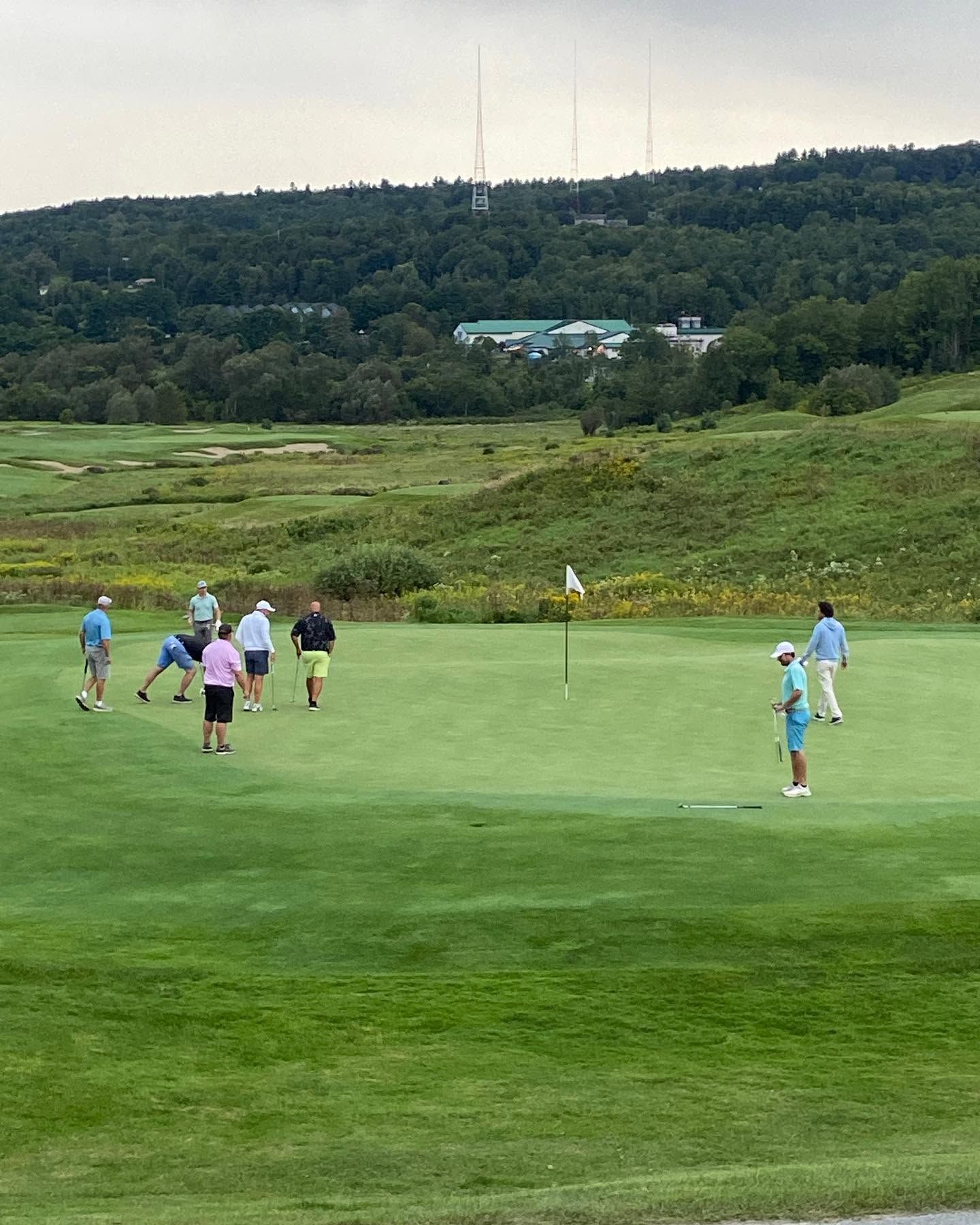 A group of golfers putt out on a large undulating green at the Country Club of Vermont in Waterbury, with rolling fescue rough, sand bunkers, and a densely forested hillside with broadcast towers visible in the background. 