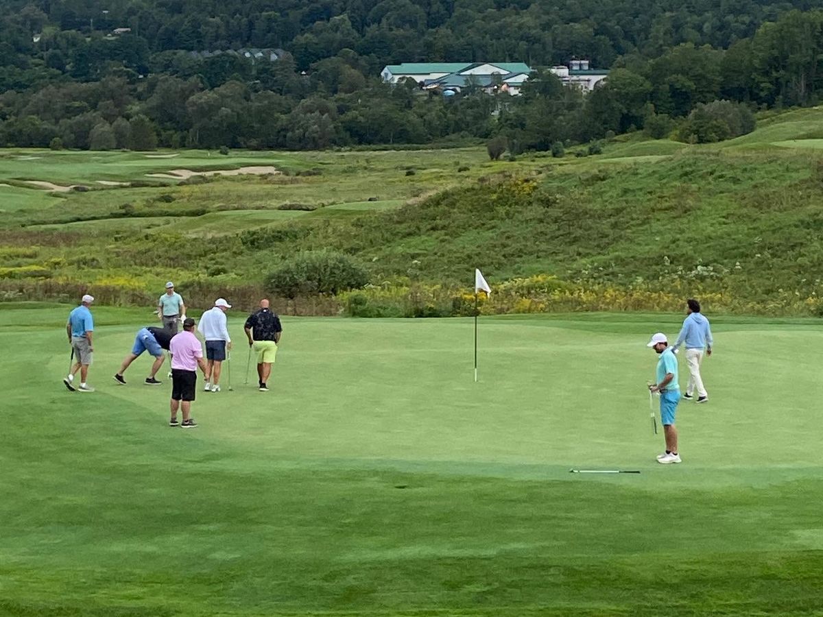 A group of golfers putt out on a large undulating green at the Country Club of Vermont in Waterbury, with rolling fescue rough, sand bunkers, and a densely forested hillside with broadcast towers visible in the background.
