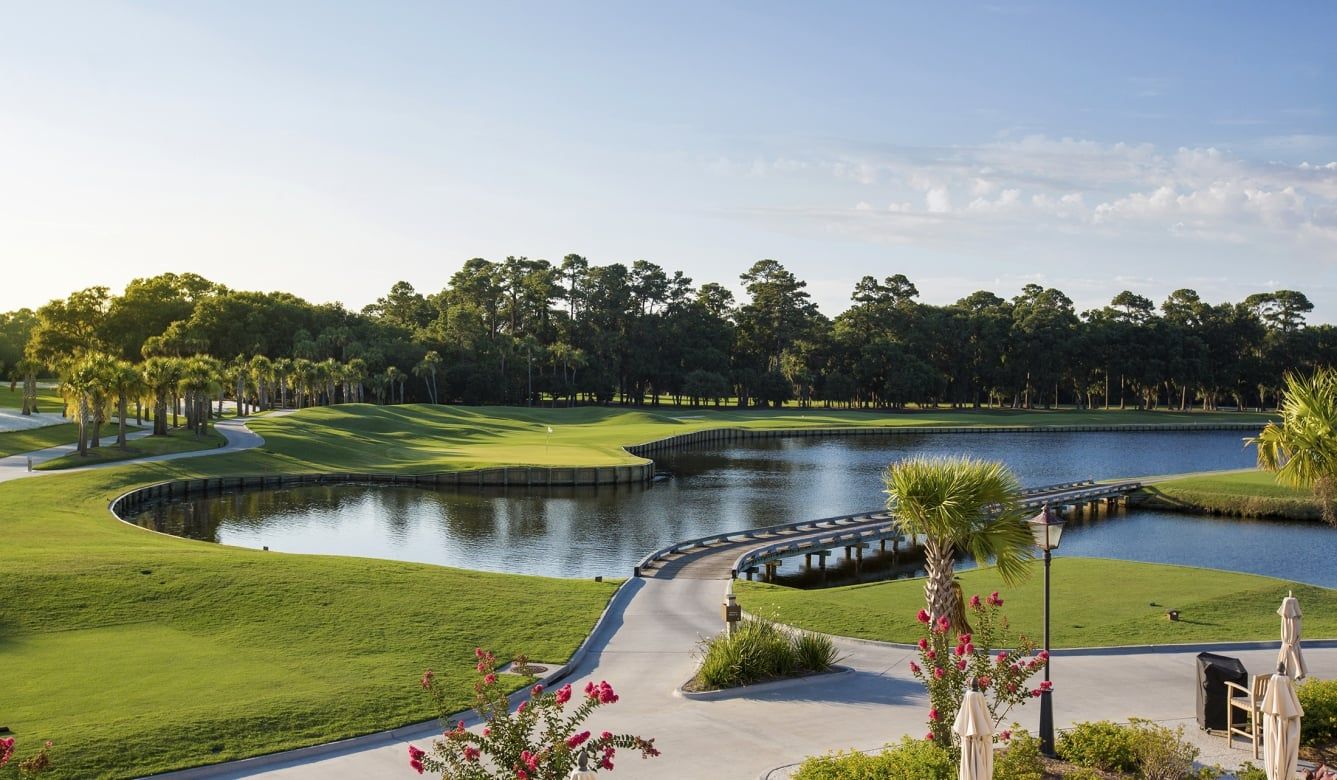 A sweeping view of Heron Point by Pete Dye on Hilton Head Island shows lush fairways winding around a serene lagoon, with a wooden footbridge, blooming crepe myrtles, and towering pines framing the course at golden hour.