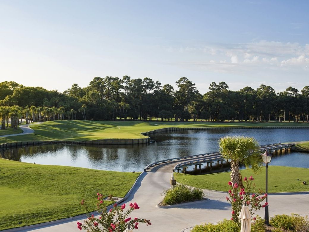 A sweeping view of Heron Point by Pete Dye on Hilton Head Island shows lush fairways winding around a serene lagoon, with a wooden footbridge, blooming crepe myrtles, and towering pines framing the course at golden hour.