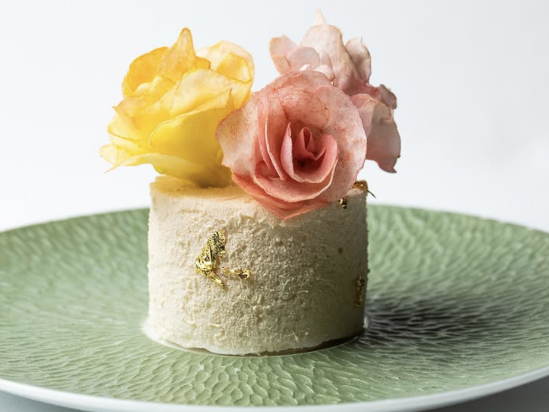 An elegant white mousse dessert adorned with delicate, lifelike yellow and pink edible flower petals and accented with gold leaf, presented on a textured green plate.