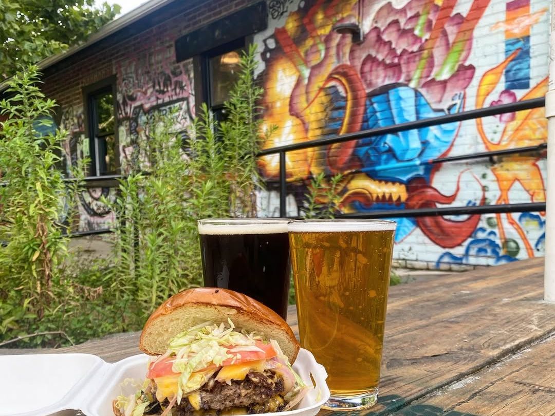 A fresh burger and two cold beers sit on a wooden table in front of a colorful mural at Wedge Brewing in Asheville. Known for its artistic setting and laid-back outdoor vibe, Wedge is a great stop for food, drinks, and street art.
