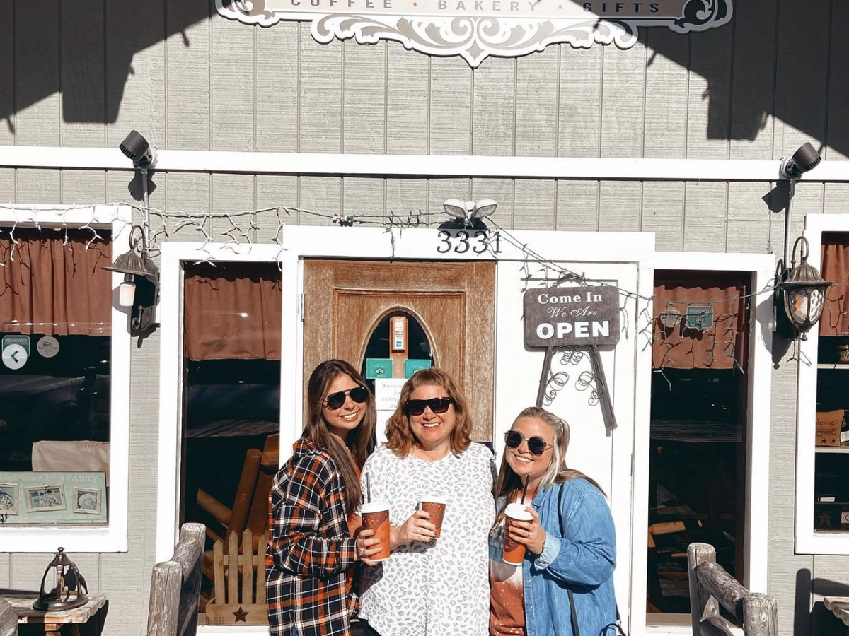 Three smiling women holding coffee drinks pose in front of a cozy café named "Espresso Yourself," with a rustic wood exterior, festive string lights, and a sign that reads "Come In, We Are OPEN."