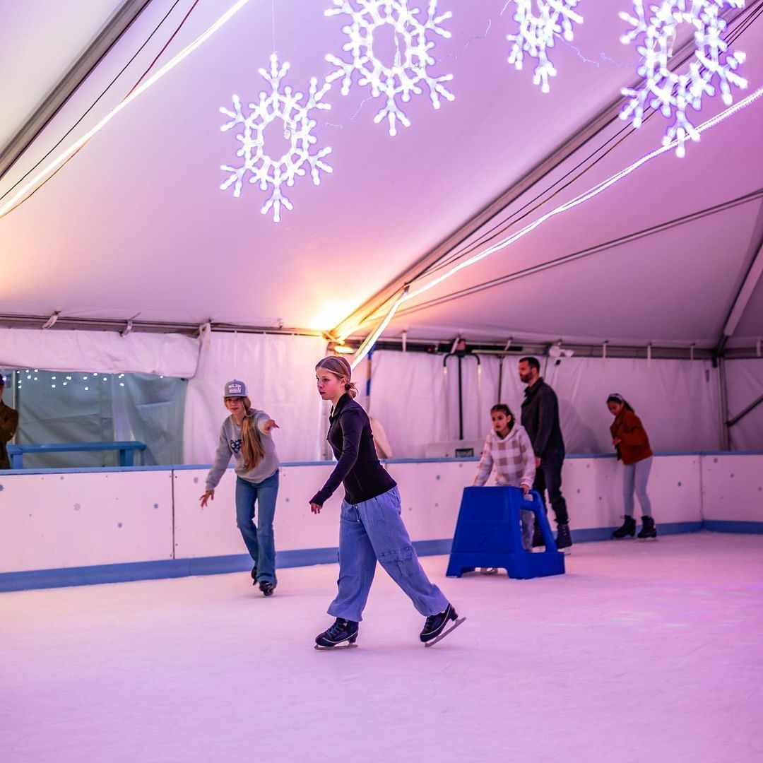 Santa Claus waves while standing on an ice rink inside a festive tent, glowing with purple lights and hanging snowflake decorations. Families skate in the background, creating a fun and cozy holiday scene perfect for winter events and Christmas celebrations. This image captures the joyful spirit of seasonal ice skating with Santa.