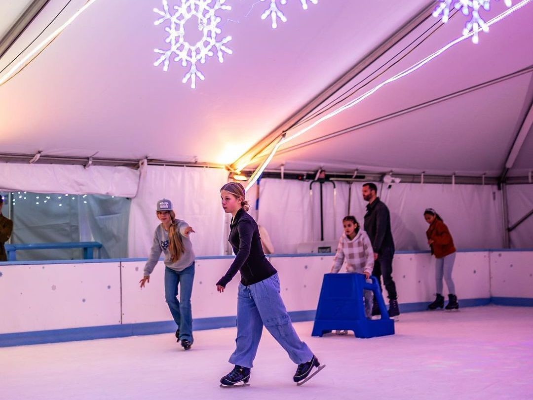 Santa Claus waves while standing on an ice rink inside a festive tent, glowing with purple lights and hanging snowflake decorations. Families skate in the background, creating a fun and cozy holiday scene perfect for winter events and Christmas celebrations. This image captures the joyful spirit of seasonal ice skating with Santa.