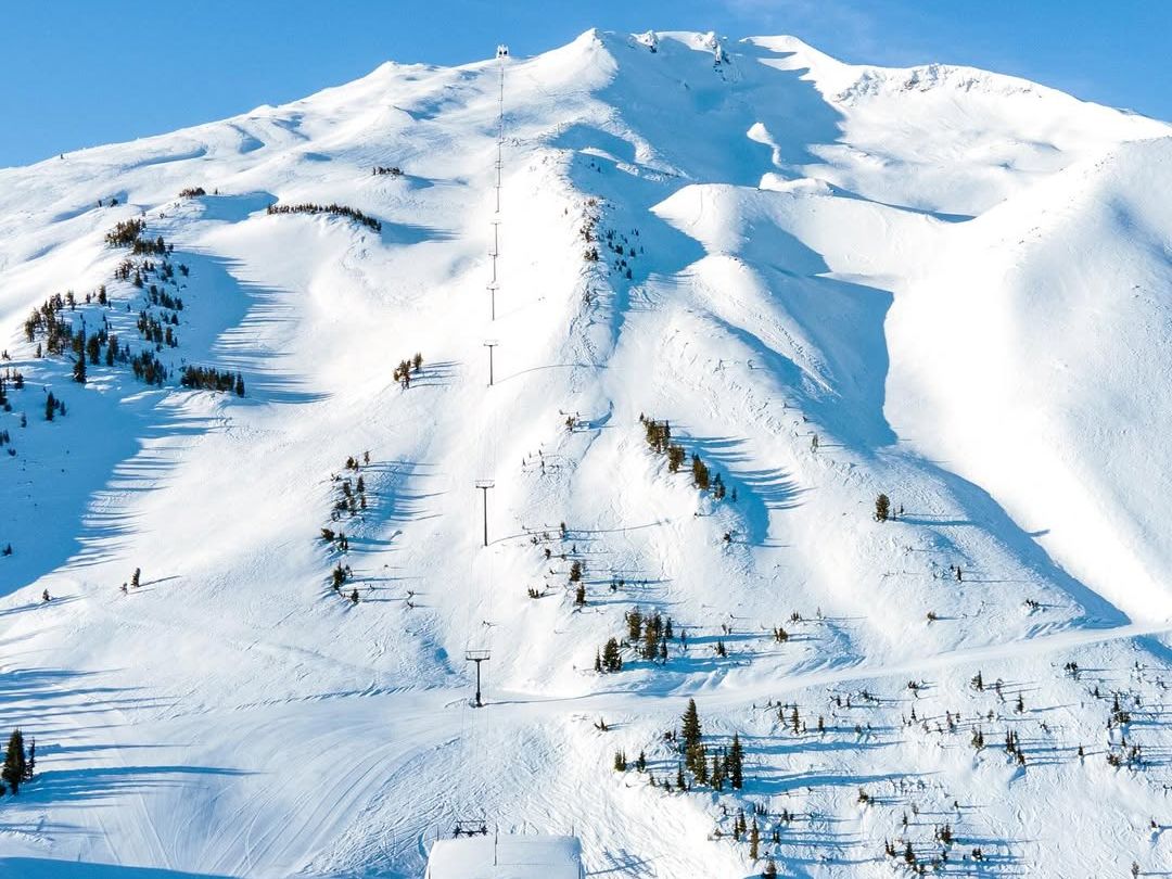 A breathtaking aerial view of Mount Bachelor covered in fresh powder, with ski lifts stretching up its slopes under a bright blue sky. A dream destination for skiers and snowboarders seeking world-class runs.