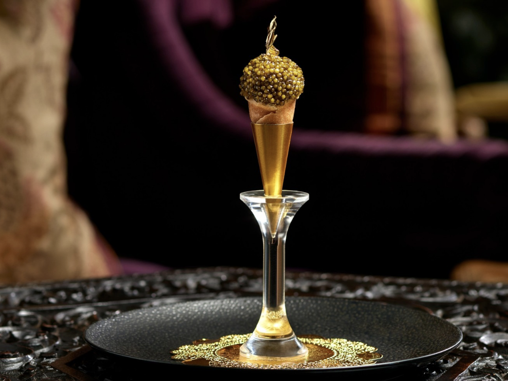 An opulent amuse-bouche featuring a mini golden cone topped with caviar and a gold leaf accent, presented elegantly on a black and gold tray with an ornate glass pedestal stand, set against a luxurious background.