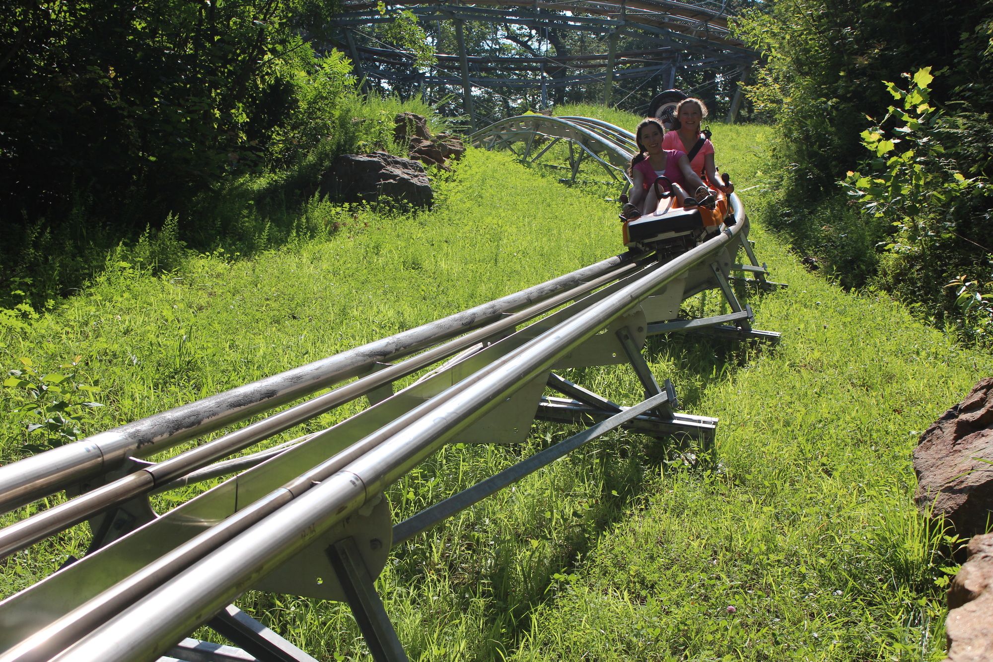 Two smiling riders glide through a green mountain trail on the Smoky Mountain Alpine Coaster. The track winds through the forest, offering a thrilling outdoor ride for families and adventure seekers.