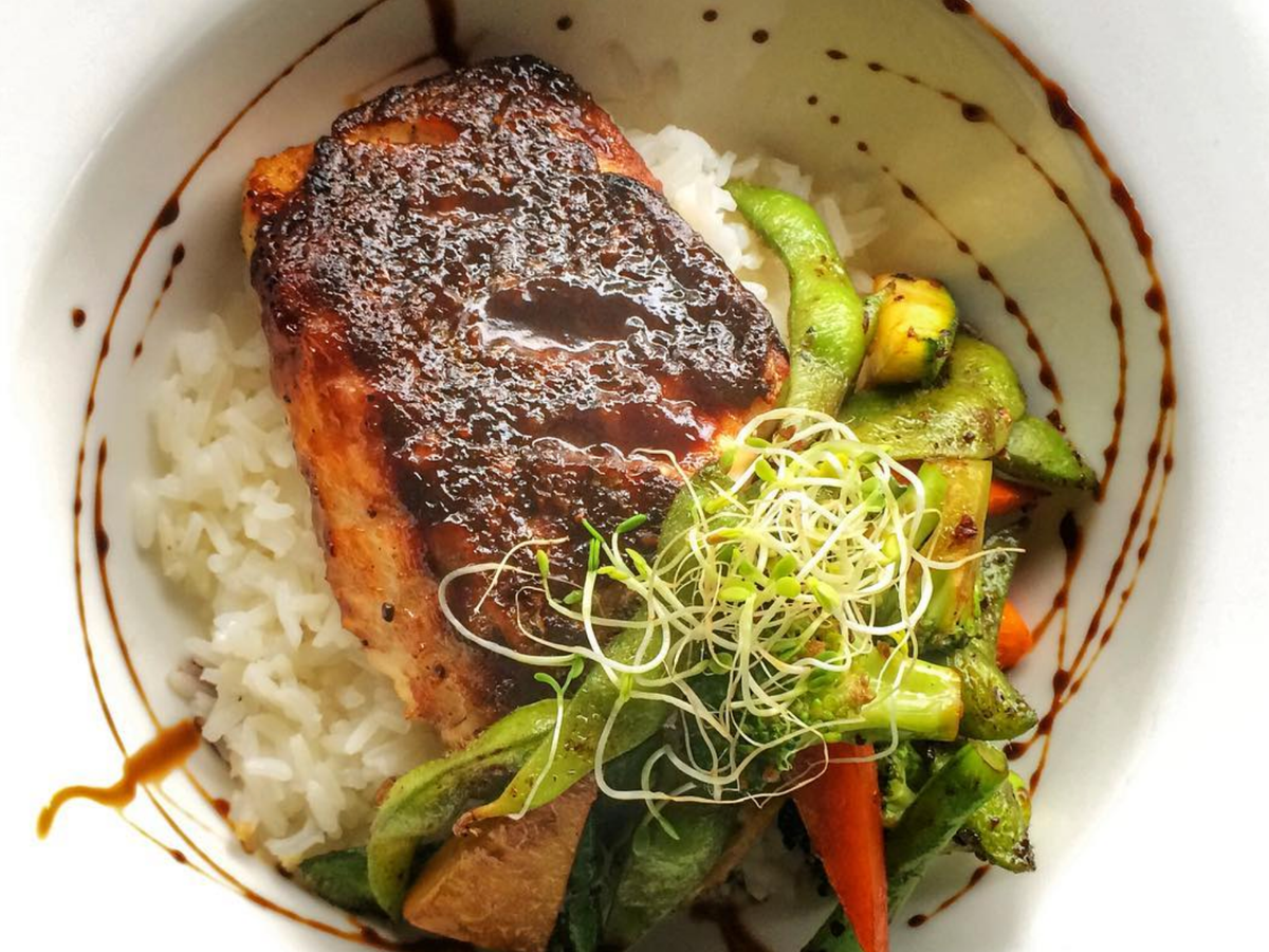 A beautifully plated dish of glazed salmon served over white rice, accompanied by sautéed vegetables and topped with fresh sprouts, all drizzled with a dark sauce in a white bowl.