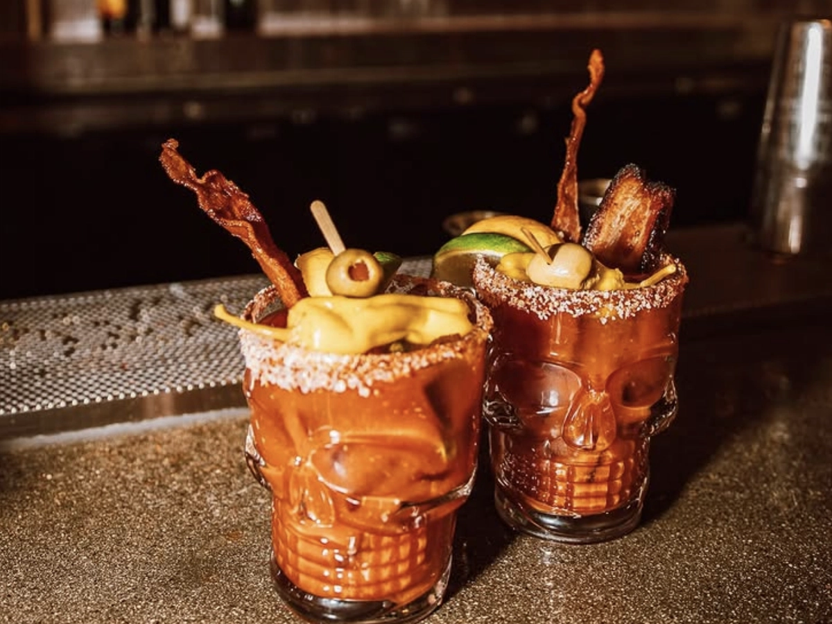 Two skull-shaped glasses filled with Bloody Mary cocktails, garnished with bacon strips, olives, lime wedges, and peppers, set on a bar counter with a salted rim.
