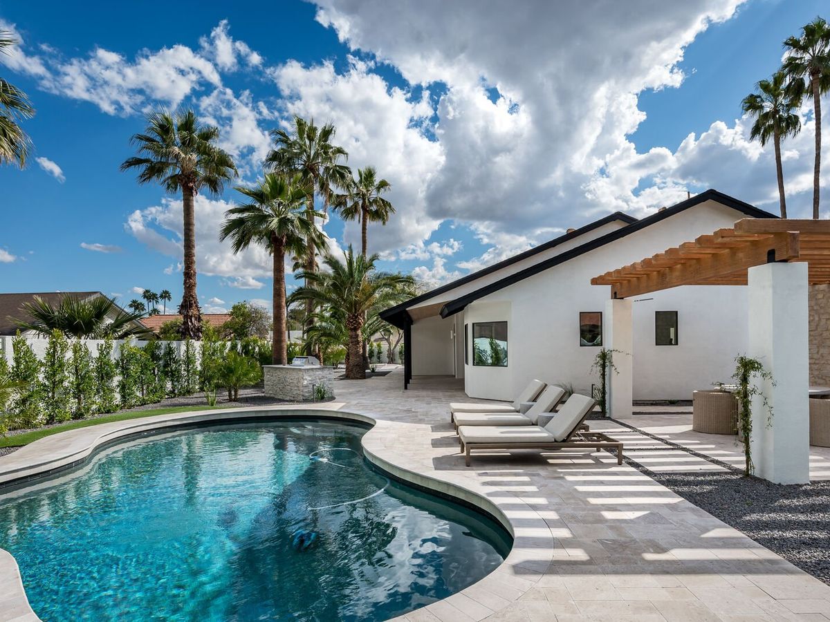 A stylish backyard features a curving swimming pool surrounded by lounge chairs, tropical palm trees, and a white modern home with a wooden pergola and outdoor seating area.