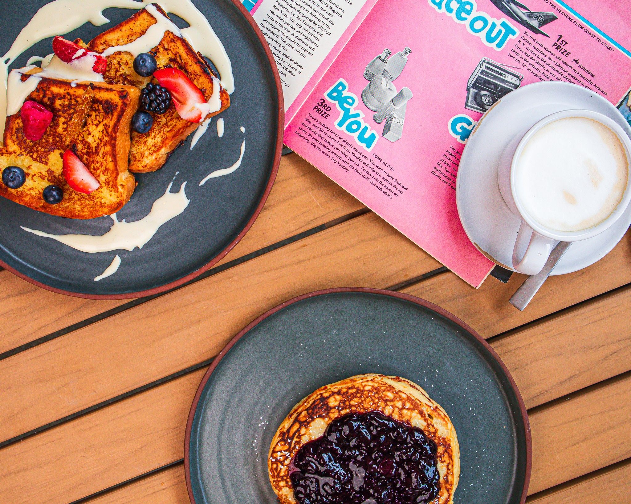 A cozy brunch scene with golden French toast, fluffy pancakes topped with fruit compote, and a steaming cup of coffee. The warm wooden table and magazine add a charming touch of everyday comfort and creativity.