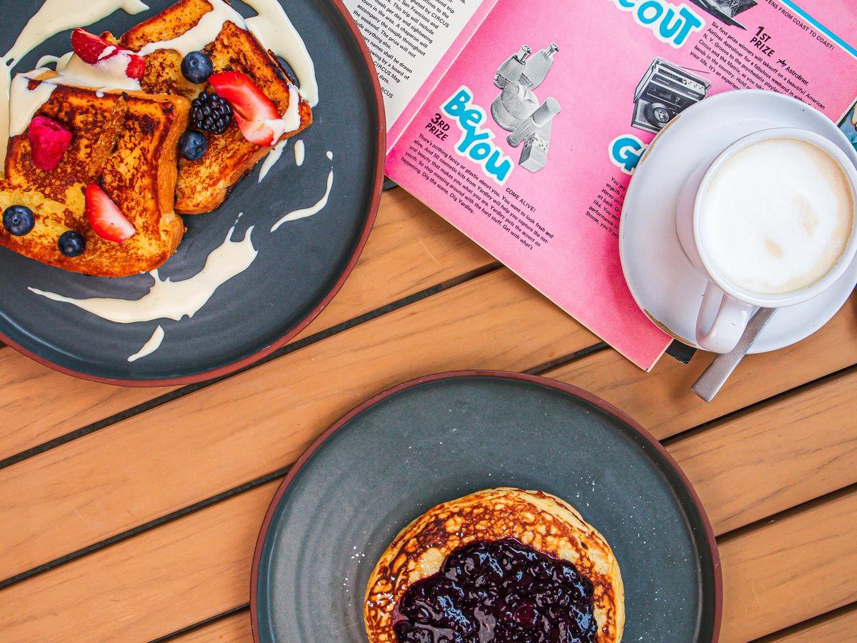 A cozy brunch scene with golden French toast, fluffy pancakes topped with fruit compote, and a steaming cup of coffee. The warm wooden table and magazine add a charming touch of everyday comfort and creativity.