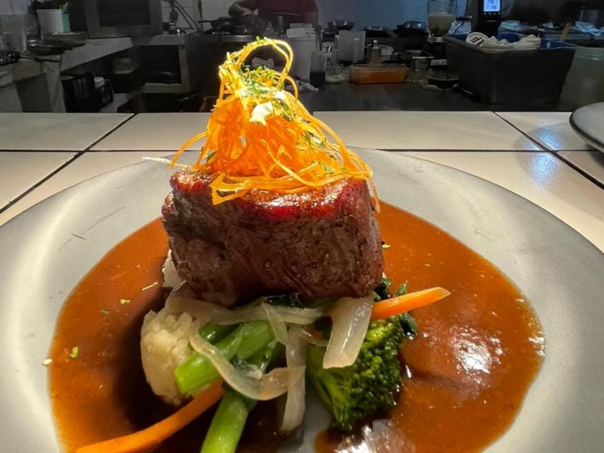 A gourmet plated dish featuring a thick-cut steak served on a bed of sautéed vegetables with a rich brown sauce, topped with a nest of crispy julienned carrots, presented in a professional kitchen setting.