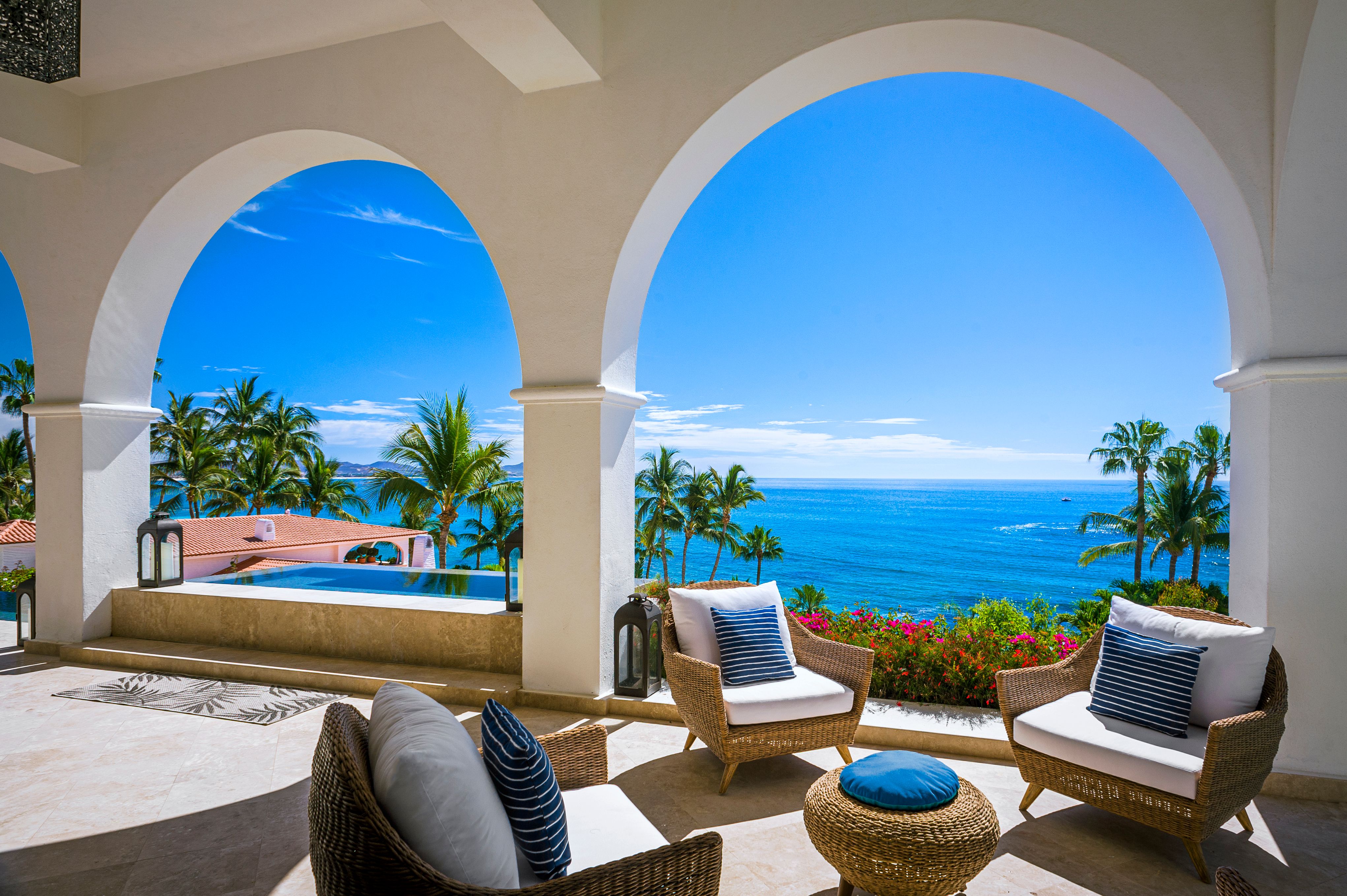 Elegant oceanfront terrace with arched columns, wicker lounge seating, and an infinity pool overlooking the turquoise sea and palm-lined coastline.