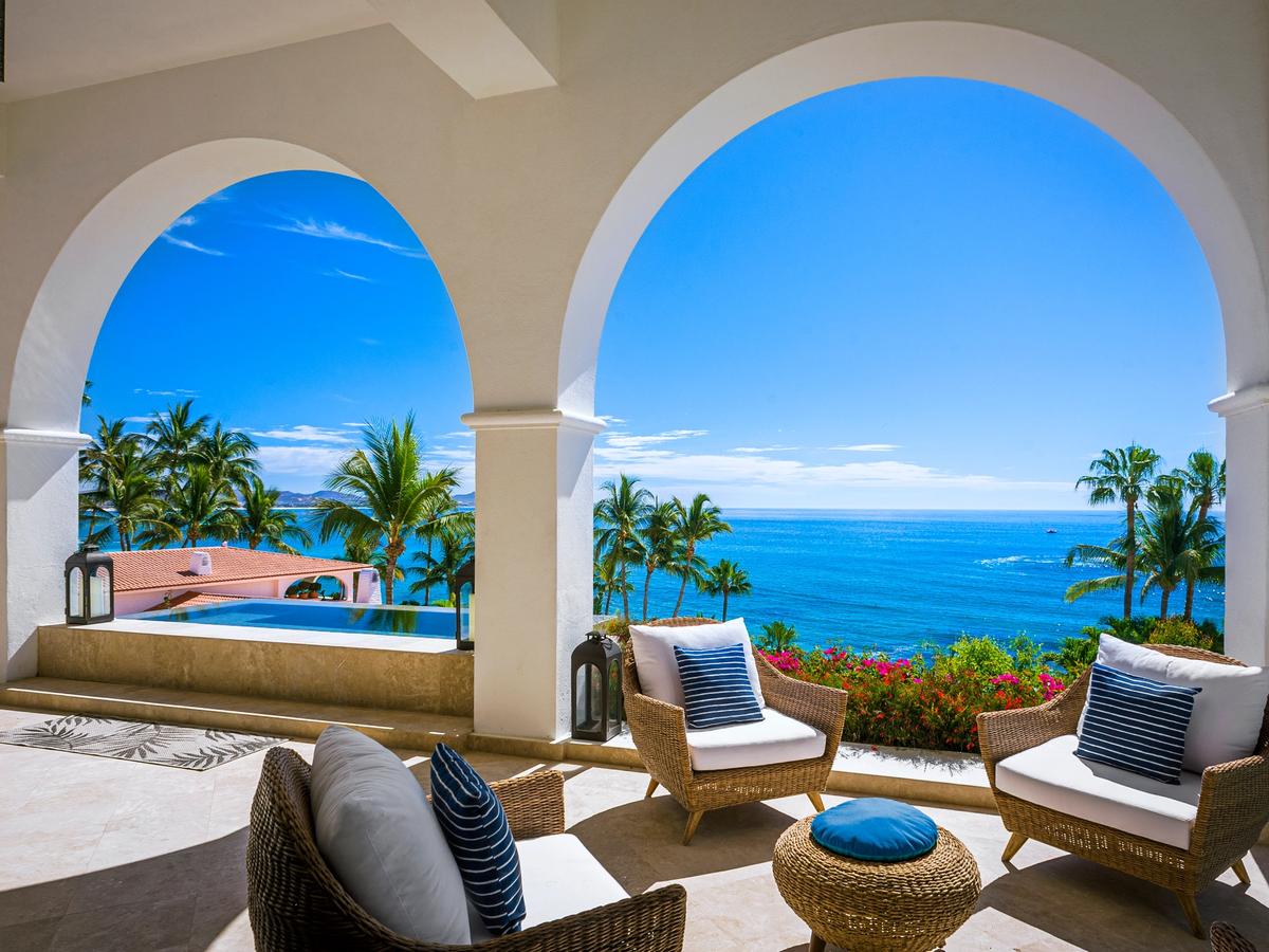 Elegant oceanfront terrace with arched columns, wicker lounge seating, and an infinity pool overlooking the turquoise sea and palm-lined coastline.