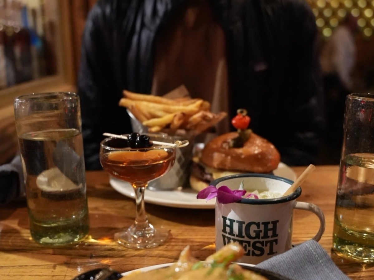 A cozy restaurant table spread with a burger and fries, two cocktails, a branded "High West" mug with a flower garnish, and a skillet of cheesy fries or poutine in the foreground.