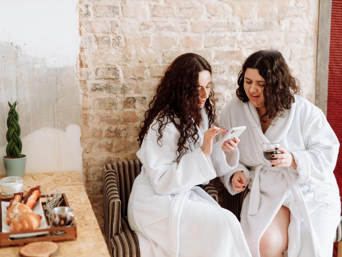 Two women in plush white robes relax side by side in striped armchairs at a spa, smiling as they look at a phone together, one holding a glass of tea. A rustic exposed brick wall, a potted plant, and a wooden tray of croissants complete the cozy, inviting setting.