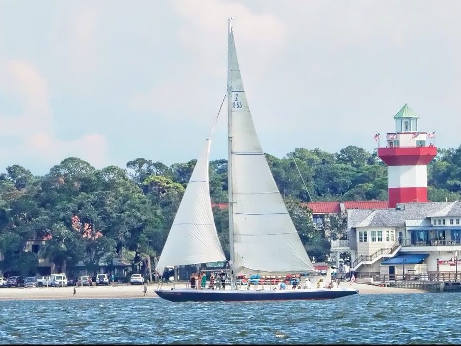 A sailboat passes the iconic Harbour Town Lighthouse during the Vagabond Sunset Dolphin Cruise in Hilton Head Island. Guests can enjoy scenic harbor views and watch dolphins play in the calm coastal waters.